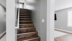 Una escalera de madera con barandilla, que conduce a una habitación con piso de tonos claros y una ventana con persianas.