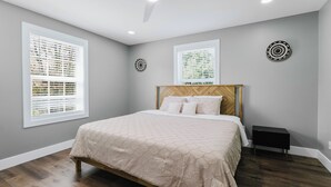 Un dormitorio con cama, dos ventanas, cabecera y mesita de noche.