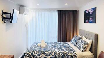 A hotel room with a bed, a TV mounted on the wall, a painting on the wall, and a window with curtains.