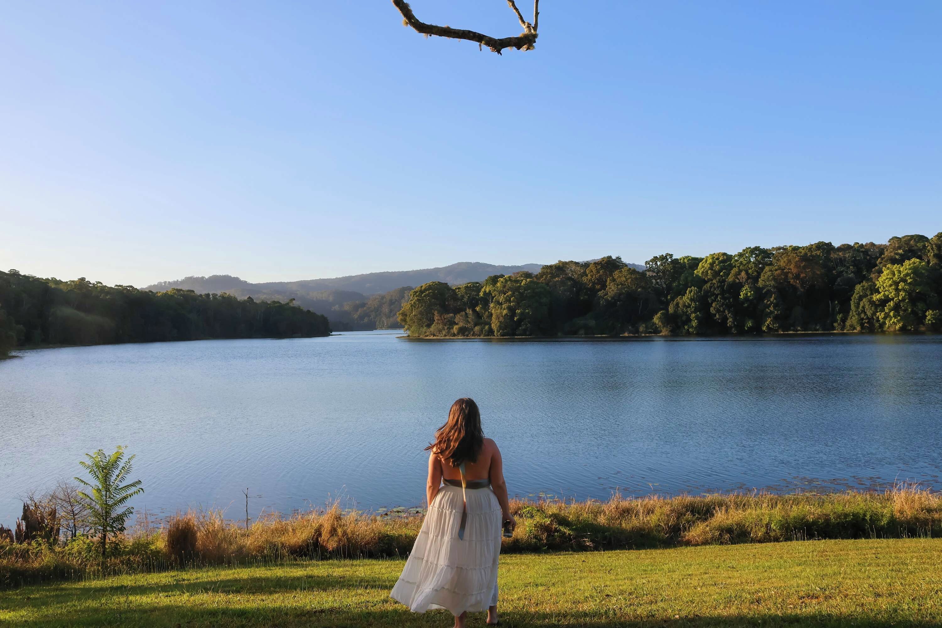 A woman in a white dress standing by a lake with a view of a forested hillside.