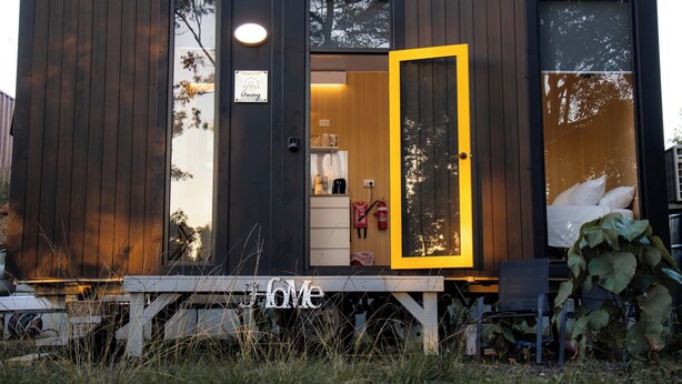 A modern cabin with a wooden exterior, a yellow door, and a visible interior with a kitchen area.