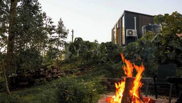 Fogueira com chamas, cercada por vegetação e um edifício ao fundo.
