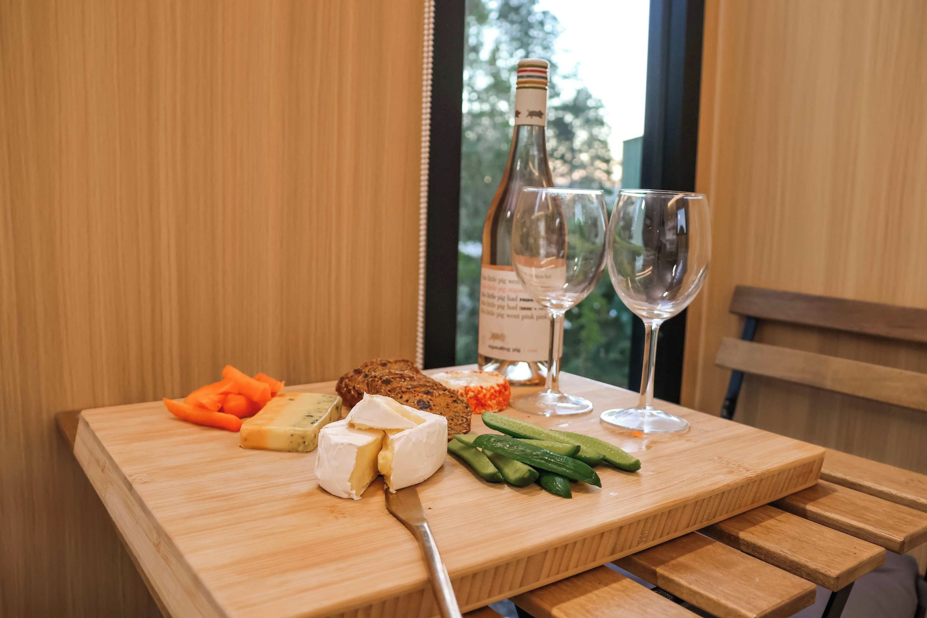 Uma mesa de madeira com uma garrafa de vinho, duas taças de vinho, um queijo, um pedaço de pão e legumes fatiados.