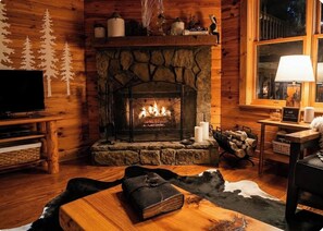 A cozy cabin interior with a stone fireplace, wooden walls, a television, and a leather-bound book on a wooden table. - Luxury Chalet Near Helen • Hot Tub, Fire Pit, Pool Table, Hammock & Creek (Clarkesville)