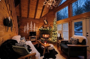 A cozy cabin interior with a fireplace, a Christmas tree, and a large window overlooking a snowy landscape.