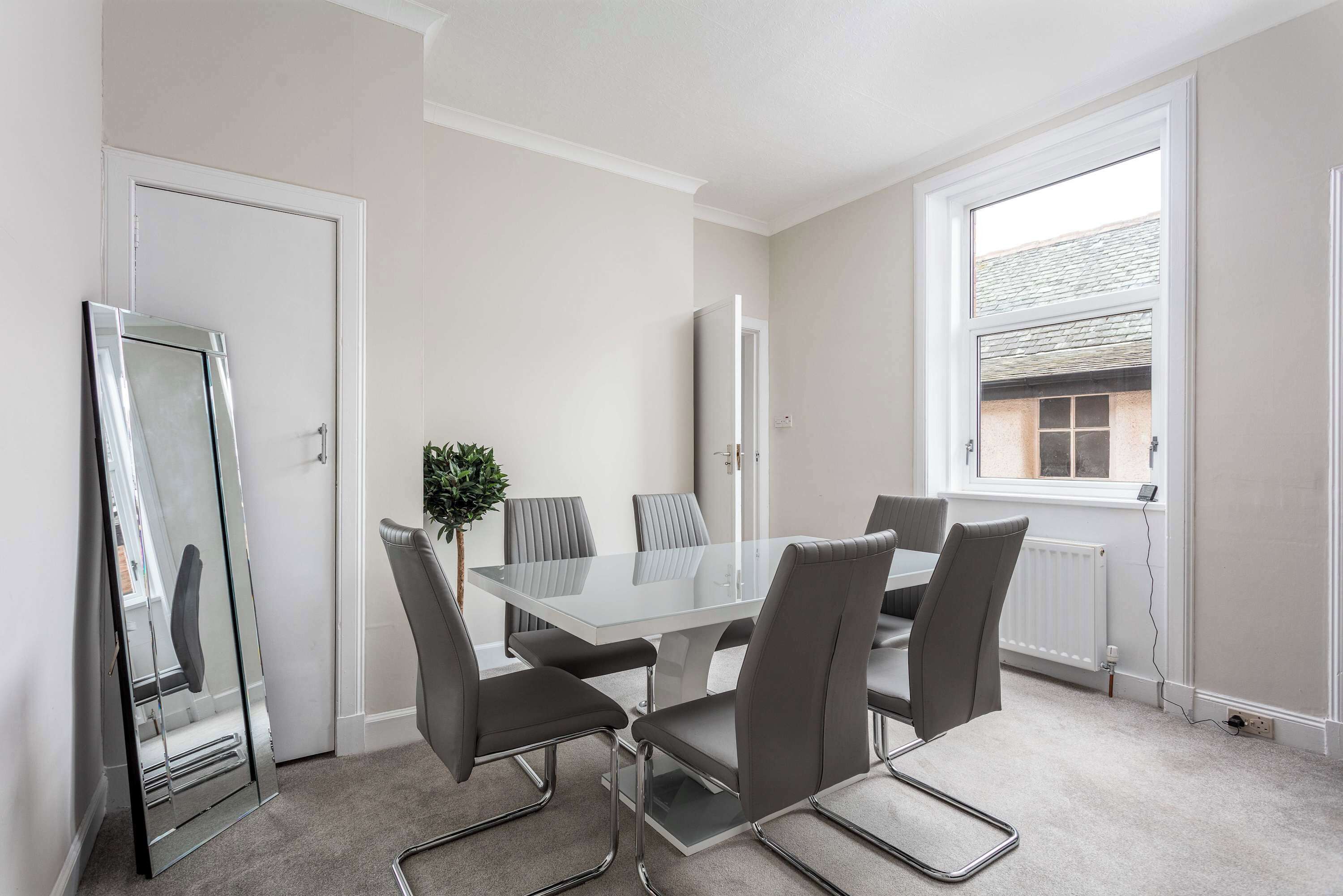 A modern meeting room with a glass table, grey upholstered chairs, a potted plant, and a view through a window.