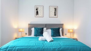 A neatly made bed with a teal bedspread and two pillows, two wall-mounted lamps, and two framed artworks above the bed. - The Bay Suite - Donnini Apartments (Troon)