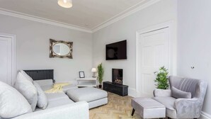 A modern living room with a grey sofa, a grey armchair, a herringbone-patterned floor, a black entertainment unit, and a large mirror on the wall. - The Bay Suite - Donnini Apartments (Troon)