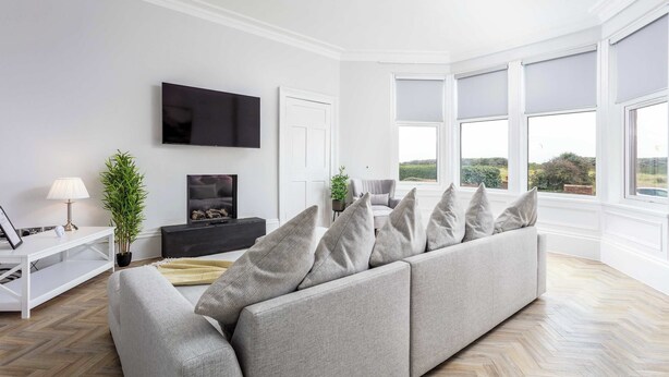 A modern living room with a grey sofa, a flat-screen TV, a white console table, and a large window with a view of greenery. - The Bay Suite - Donnini Apartments (Troon)