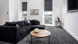 Un salon moderne avec un canapé d’angle noir, une table basse ronde en bois et une vue sur la ville grâce à de grandes fenêtres.