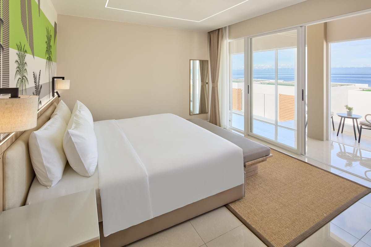 Ett hotellrum med en stor säng, ett skrivbord med en lampa, utsikt över stranden och en balkong med ett bord och stolar.
