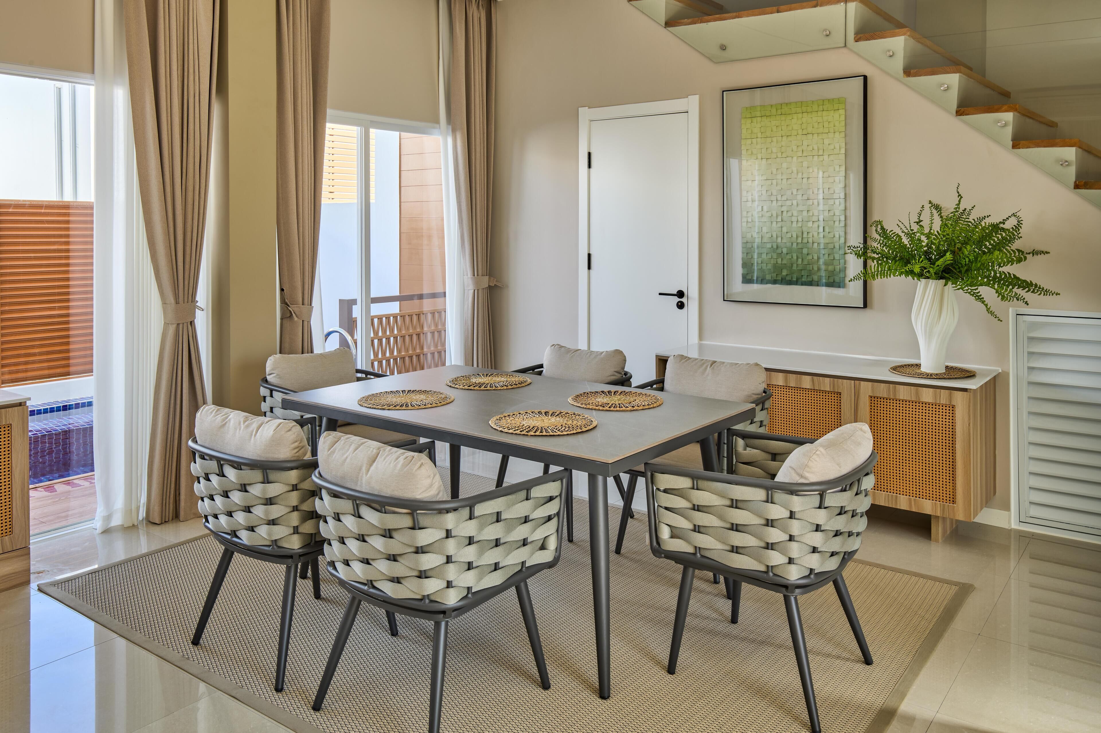 Comedor con una mesa lista para cuatro personas, rodeada de sillas de mimbre con cojines.