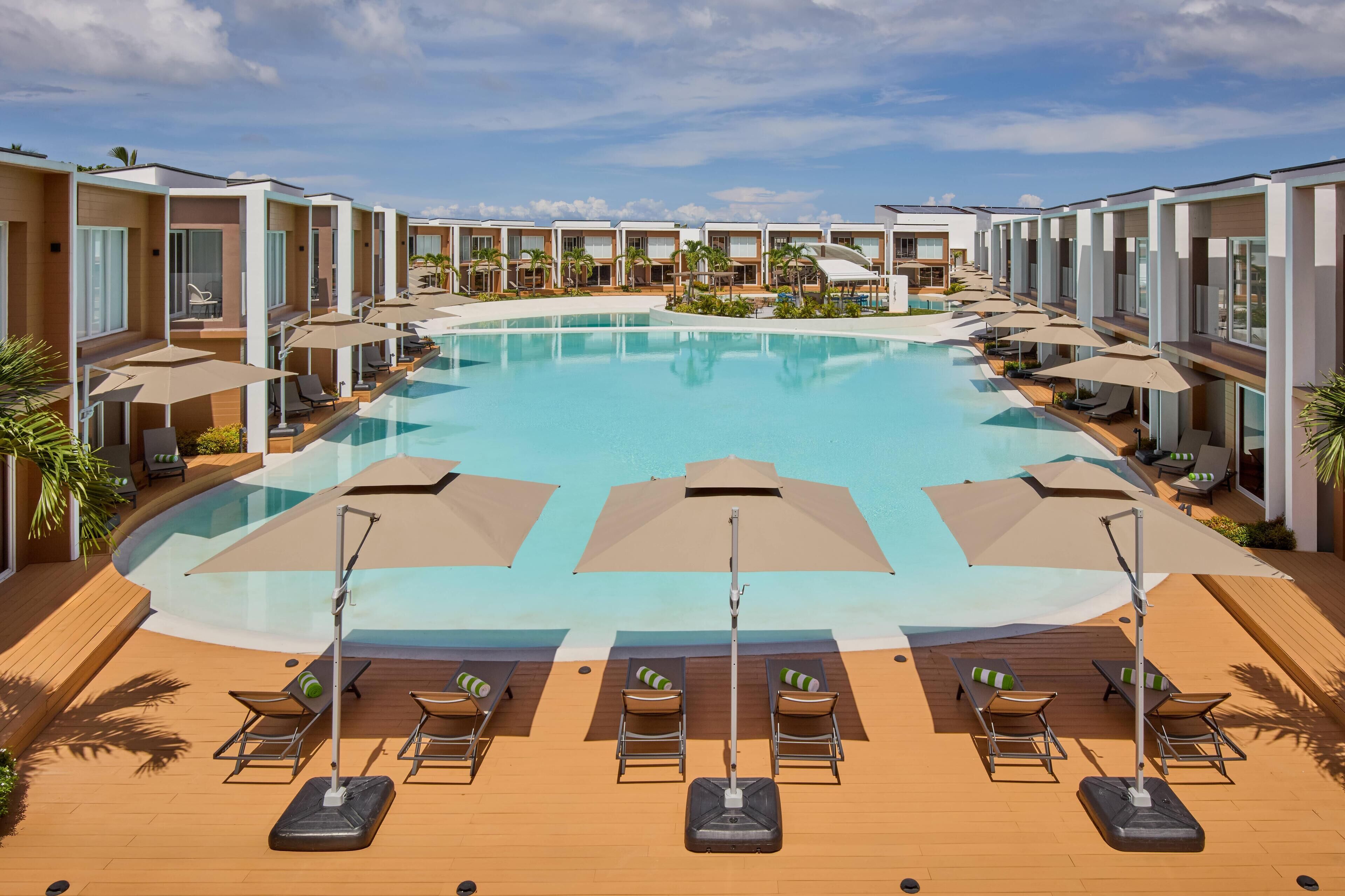 A large swimming pool surrounded by modern buildings with outdoor seating areas and umbrellas.