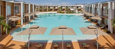A large swimming pool surrounded by modern buildings with outdoor seating areas and umbrellas.