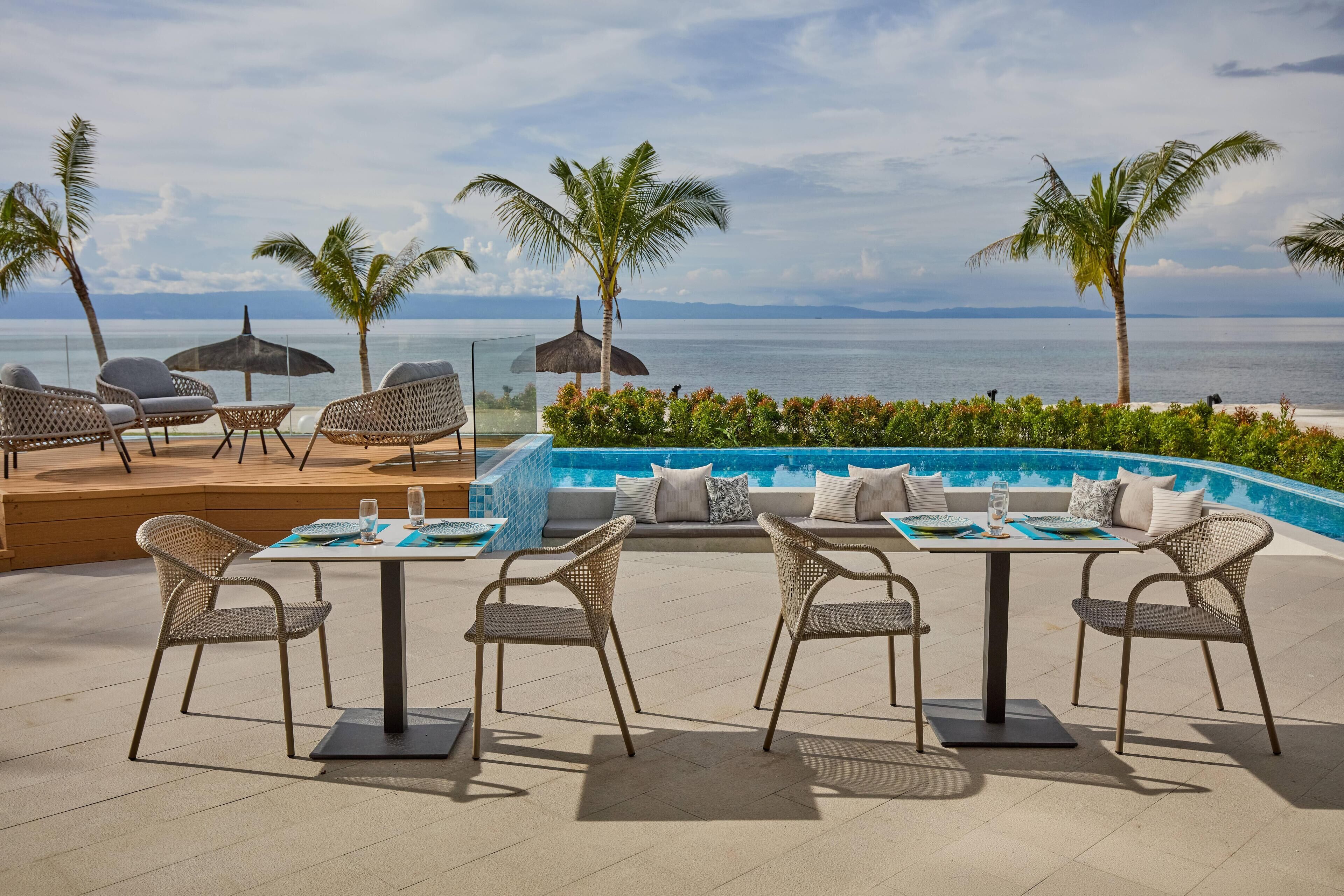 A poolside area with wicker chairs and tables, a seating area with cushions, and a view of the ocean.