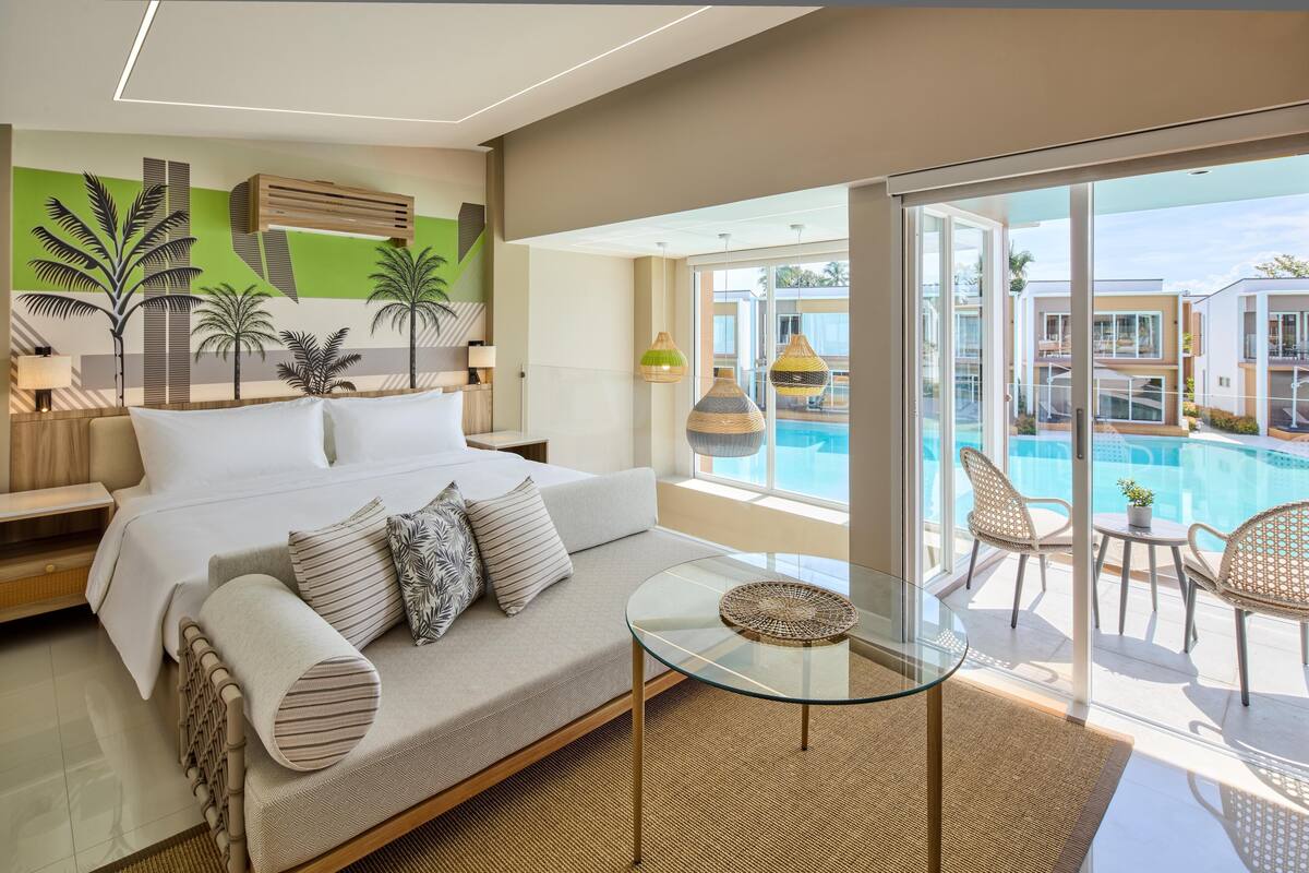Ett modernt hotellrum med en stor säng, ett soffbord i glas, en soffa och utsikt över en swimmingpool och andra byggnader.