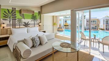 A modern hotel room with a large bed, a glass coffee table, a sofa, and a view of a swimming pool and other buildings.