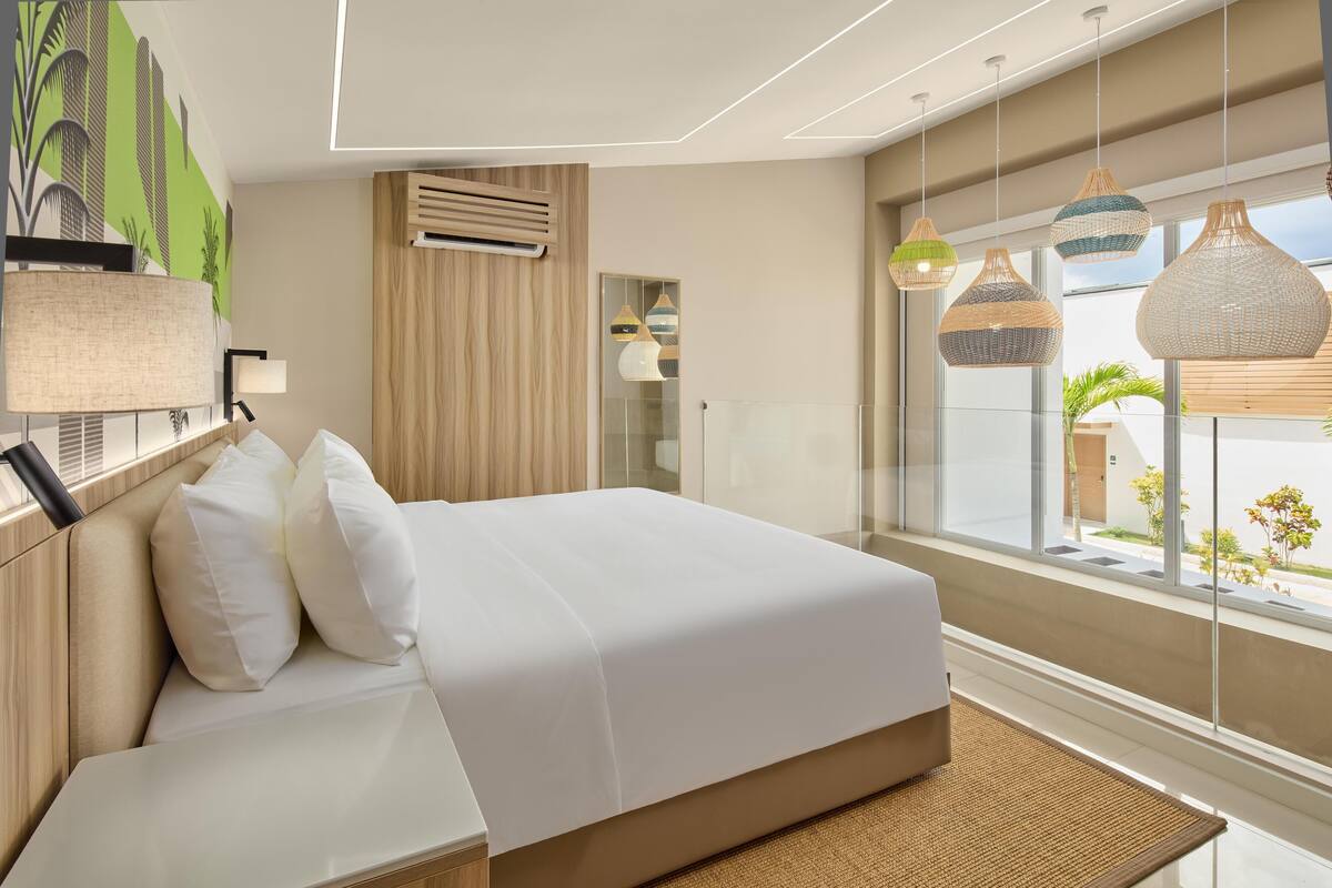 Ett modernt hotellrum med en stor säng, utsikt över staden och ett stort fönster med hängande lampor.