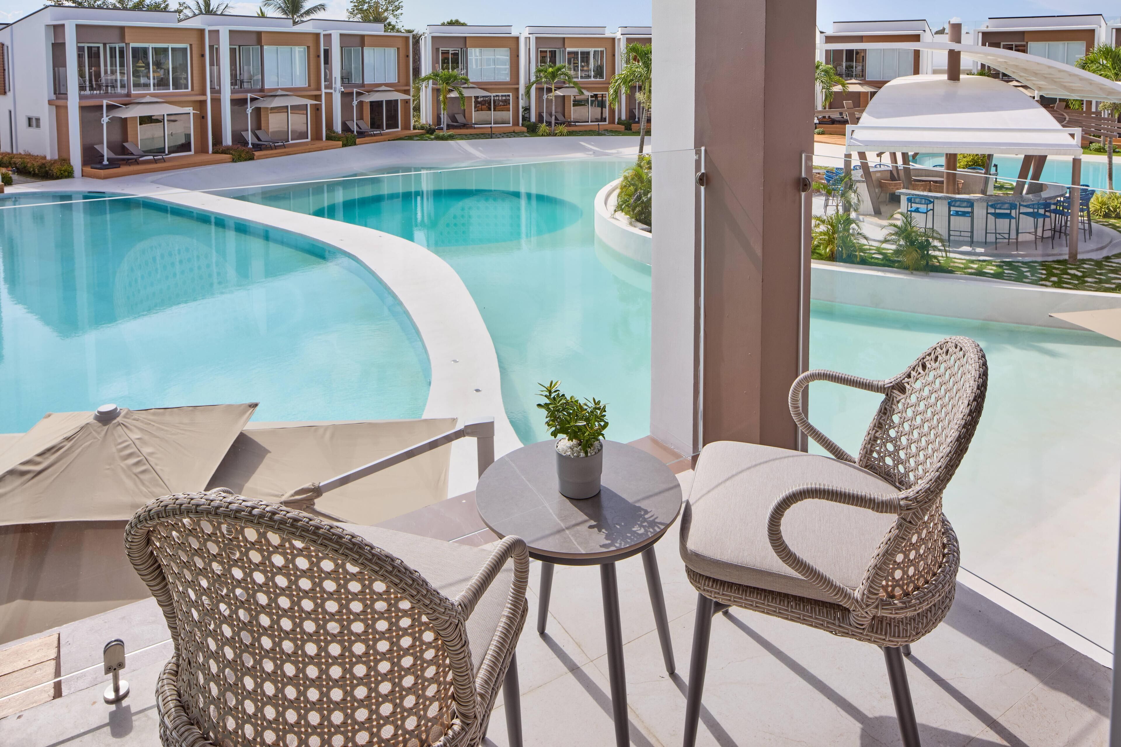 Zona de piscina con sillas de mimbre y una pequeña mesa, con vista a una piscina grande y edificios cercanos.