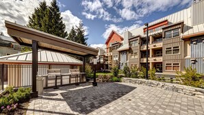 A courtyard with a barbecue area, stone paving, and a building with multiple balconies and a red roof. - Lake Placid Lodge - Ski-In/Out, Pool, Hot Tub & Sauna, No Stairs (Whistler)