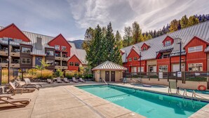 A pool area with lounge chairs, a small building, and red-roofed buildings in the background. - Lake Placid Lodge - Ski-In/Out, Pool, Hot Tub & Sauna, No Stairs (Whistler)