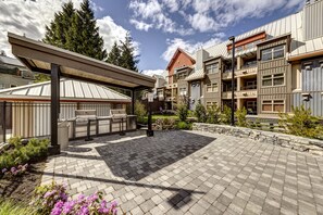 A covered outdoor barbecue area with a grill and recycling bin, surrounded by a paved patio and stone walls. - Lake Placid Lodge 126 (Whistler)