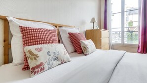 Una cama bien hecha con ropa blanca y varios almohadones decorativos en una habitación con cabecera de madera, una mesita de noche con lámpara y una ventana con cortinas moradas.