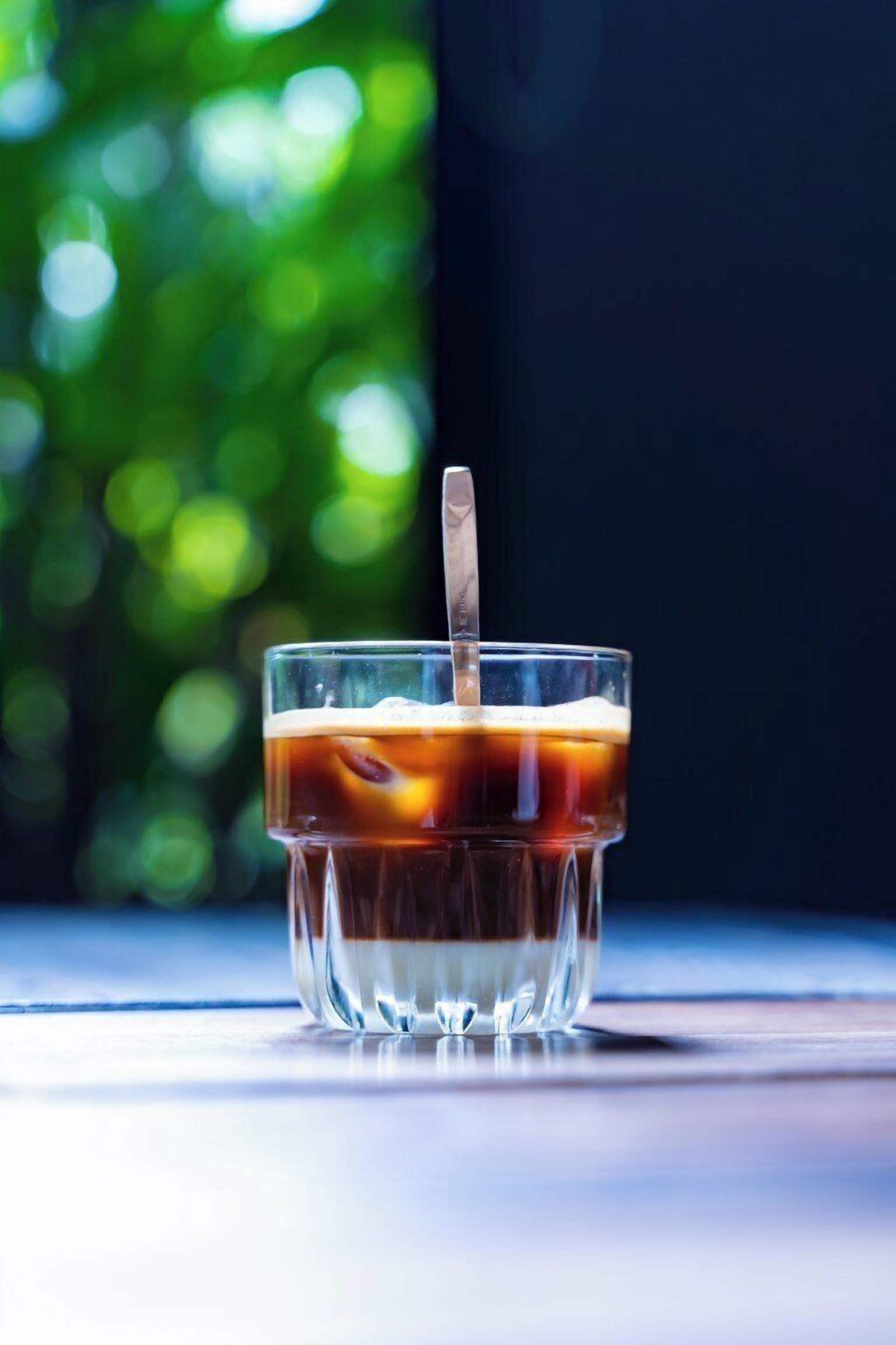 A clear glass with a straw, containing a dark liquid, possibly iced tea, on a reflective surface with a blurred green background.