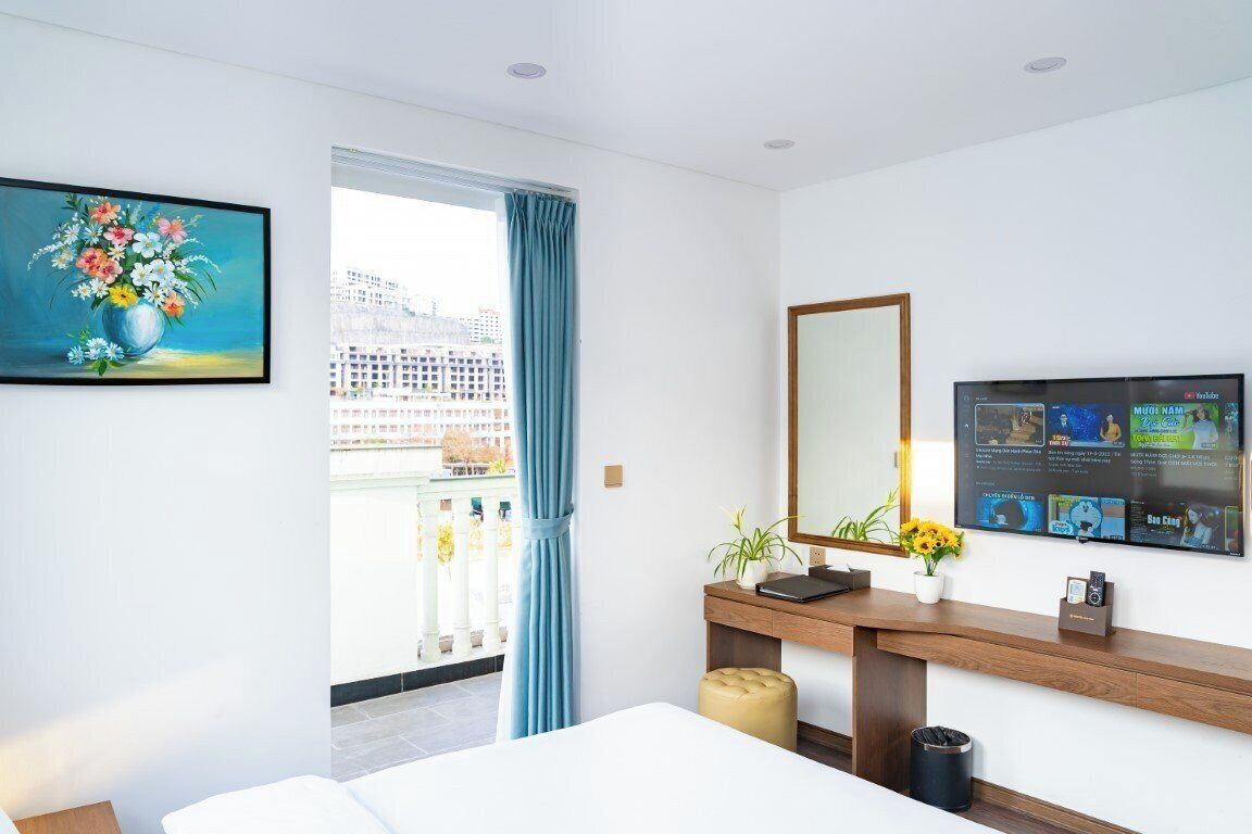 Hotellihuone, jossa on sänky, työpöytä tietokoneella, televisio, ikkuna verhoilla ja taulu seinällä.