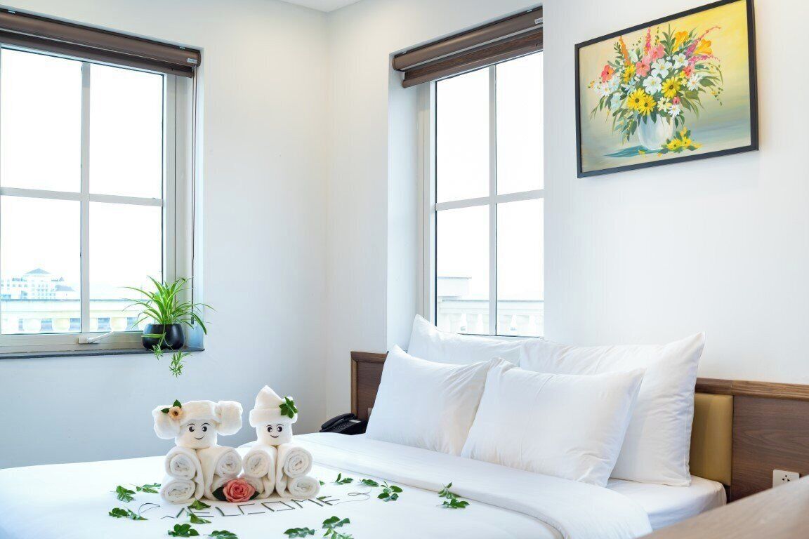 A hotel room with a large bed, two windows, a painting of flowers, and a small potted plant.