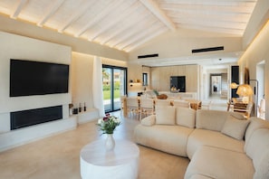 A modern living room with a large flat-screen TV, a sectional sofa, and a dining area with a kitchen in the background.