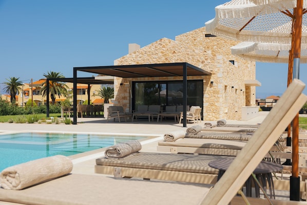 A poolside area with lounge chairs, a covered seating area, and a stone building with a glass enclosure.
