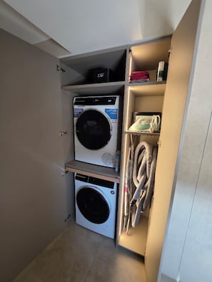 A compact laundry room with two washing machines and a shelf with towels and a red box.