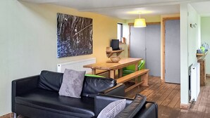 A living room with a black leather sofa, a wooden table, and a green chair. There is a radiator, a painting on the wall, and a door leading to a kitchen area.