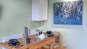 A wooden dining table with a kettle, cups, and a coffee pot. A bench is attached to the table. A chair is visible.