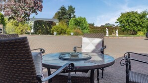 Un espace patio aménagé avec des chaises en osier et une table en verre, entouré d’un chemin en briques et de verdure.