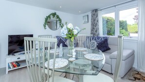 Un salon moderne avec une table à manger en verre, des chaises blanches, un canapé agrémenté de coussins à motifs, un téléviseur et une grande fenêtre avec des rideaux.