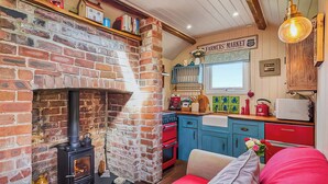 Una sala de estar acogedora con chimenea de ladrillo, un sofá rojo y una zona de cocina con armarios azules y un hervidor rojo.