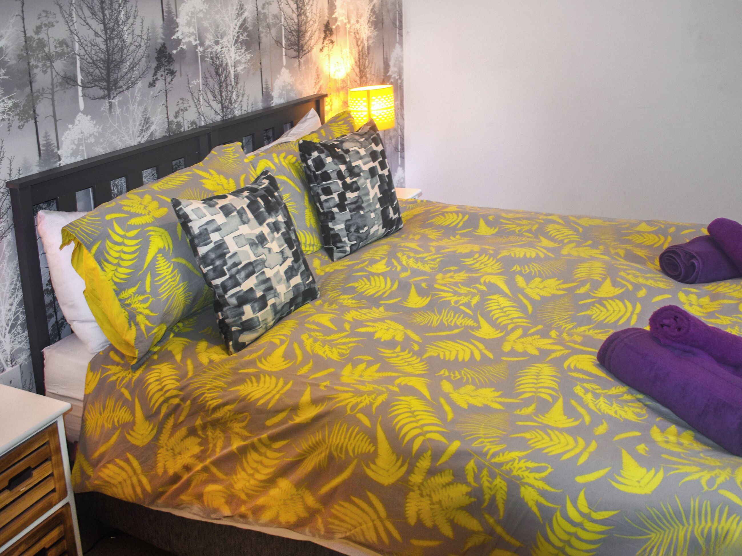 A bed with a patterned bedspread, pillows, and a headboard against a wall with a tree mural.
