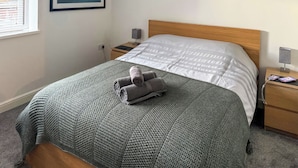 Un letto rifatto con cura, una coperta fantasia, asciugamani arrotolati, un comodino con una lampada, un quadro appeso al muro e una finestra con persiane.