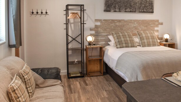 Un dormitorio con cama, mesita de noche, suelo de madera, estante en la pared y ventana con cortinas.