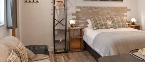 A bedroom with a bed, a nightstand, a wooden floor, a wall-mounted shelf, and a window with curtains.