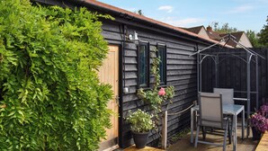 Una casa de madera con exterior oscuro, un muelle de madera, una silla metálica y plantas en macetas.