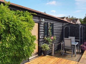 Une maison en bois à l’extérieur sombre, avec une terrasse en bois, une chaise en métal et des plantes en pot.