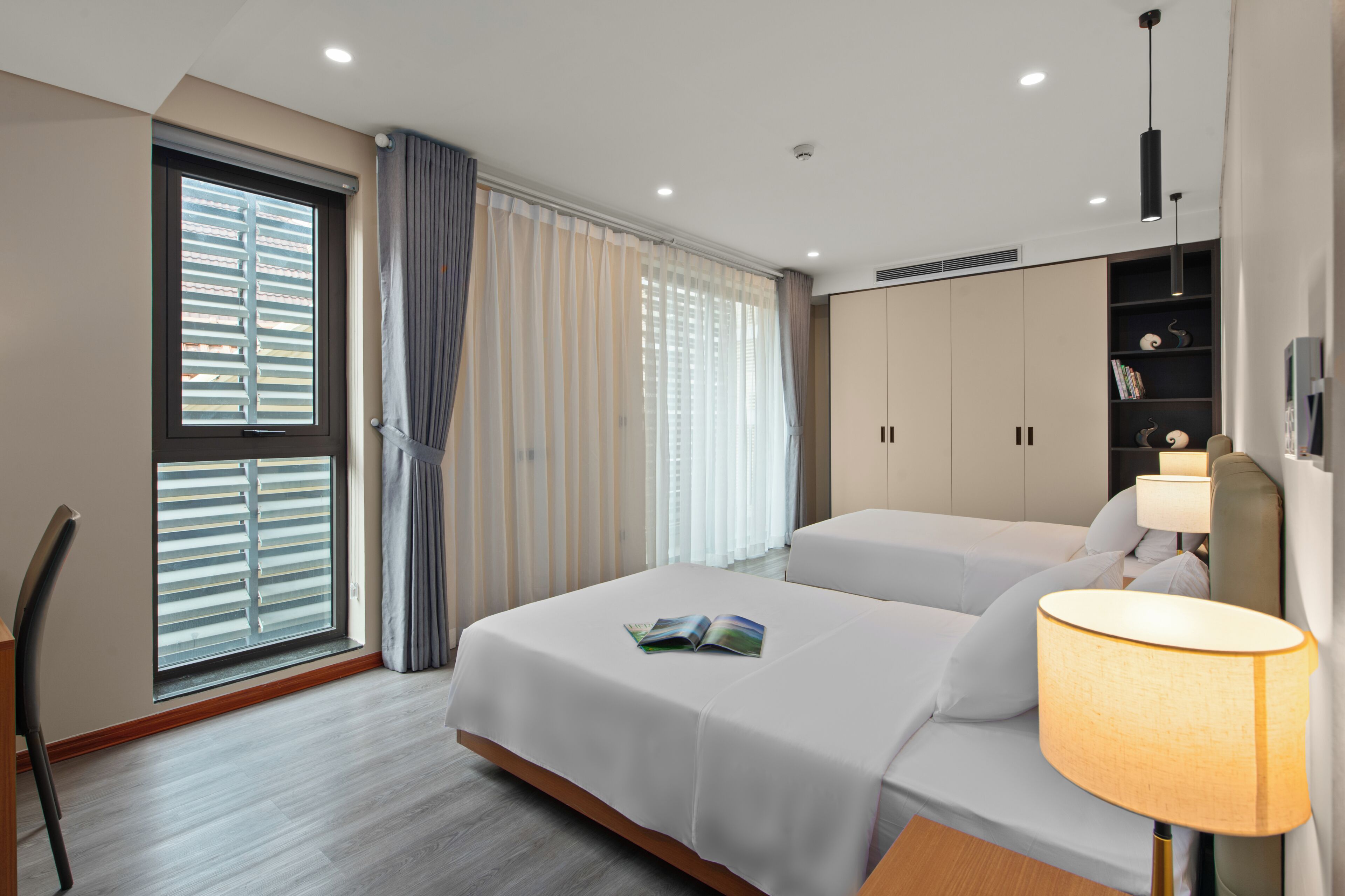 A hotel room with two beds, a desk, a chair, a window with blinds, a lamp, and a wardrobe.