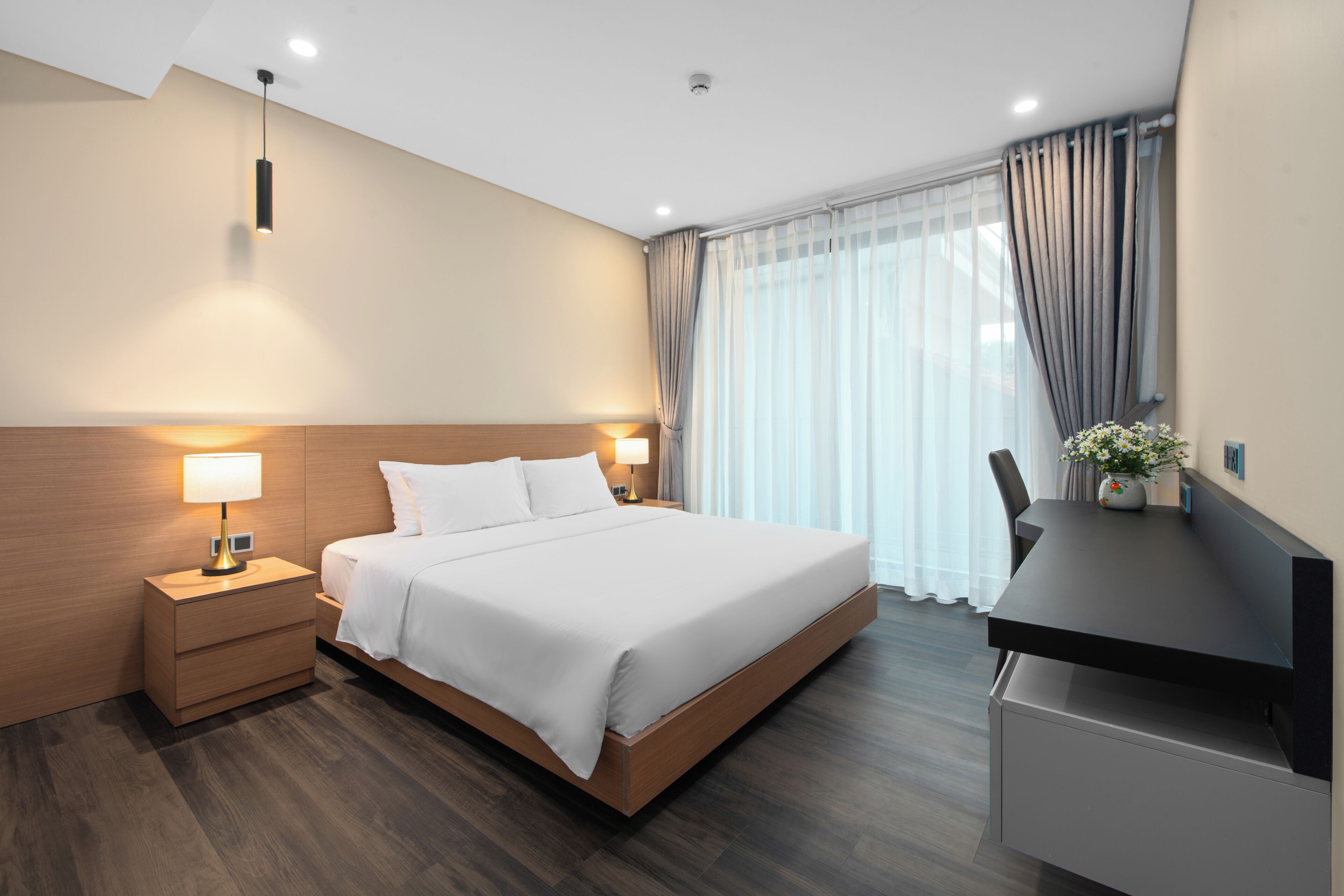 Ein modernes Hotelzimmer mit einem Holzkopfteil, einem großen Bett, Nachttischlampen, einem Schreibtisch mit Stuhl und einem Fenster mit Vorhängen.