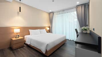 A modern hotel room with a wooden headboard, a large bed, bedside lamps, a desk with a chair, and a window with curtains.