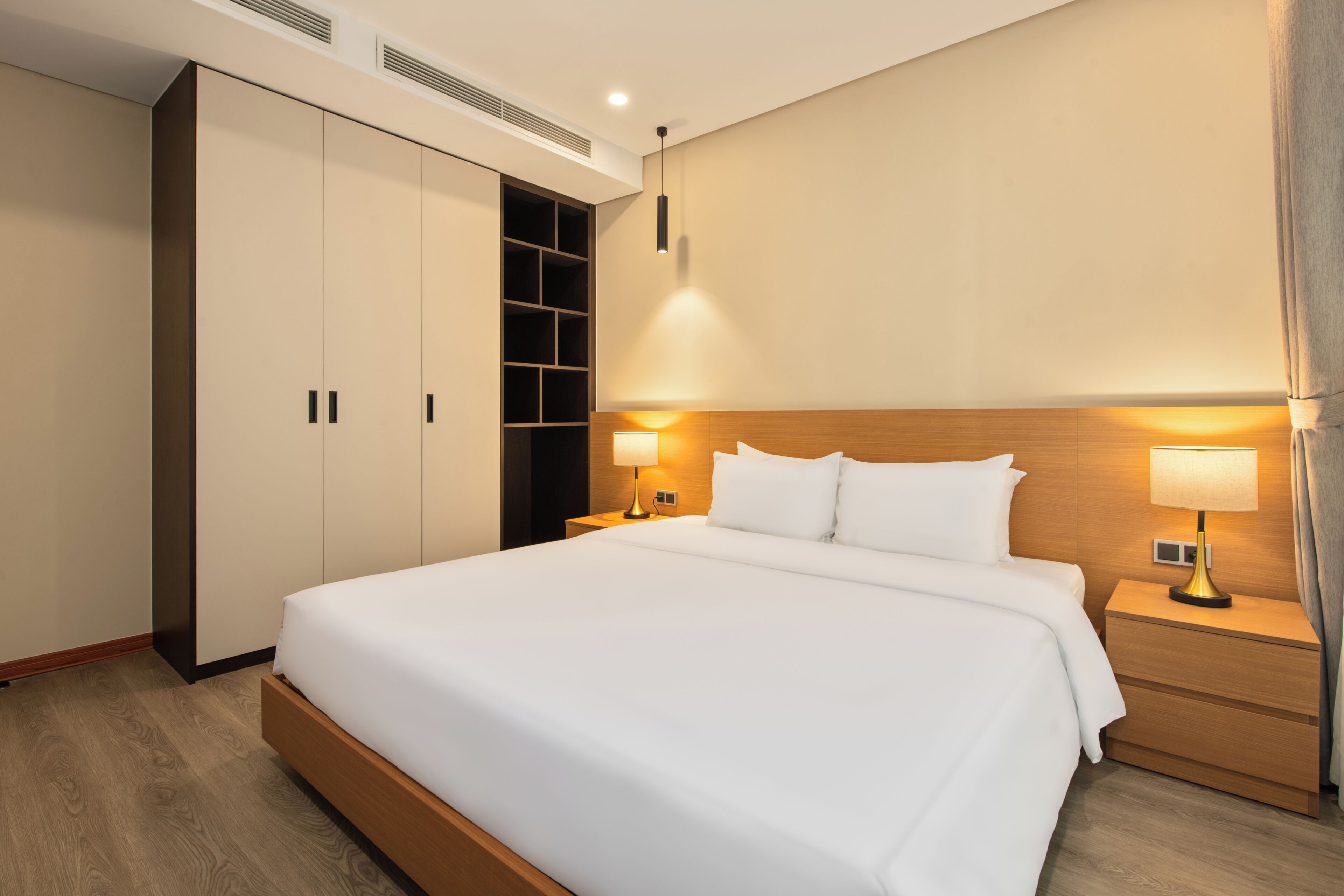 A hotel room with a large bed, two bedside tables with lamps, a wardrobe, and a built-in shelf unit.