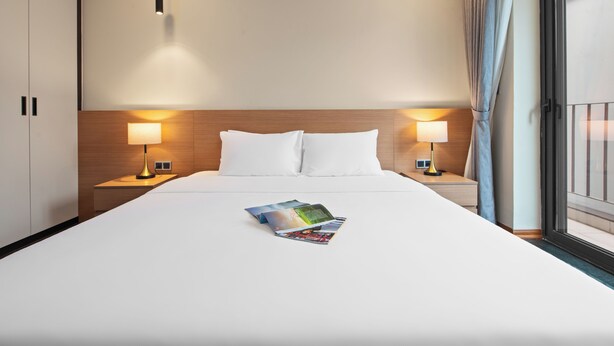 A hotel room with a large bed, two bedside lamps, a wooden headboard, and a window with curtains. - Regal Residences Tay ho (Hanoi)
