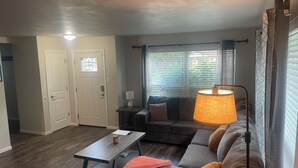 A living room with a sofa, a coffee table, a lamp, and a window with blinds. - Charming 2-bedroom house in Jefferson City with AC (Jefferson City)